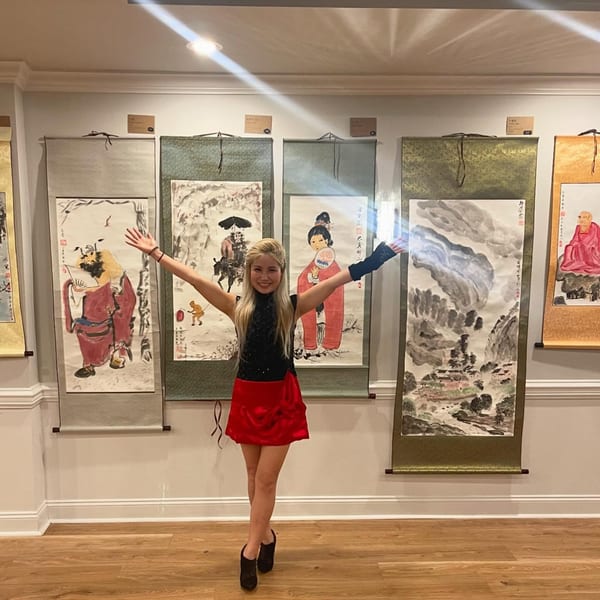 A woman in a black top and red skirt stands with arms outstretched in front of Asian art scrolls.