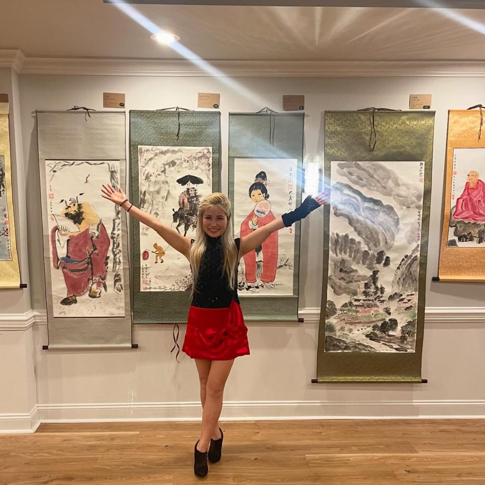 A woman in a black top and red skirt stands with arms outstretched in front of Asian art scrolls.