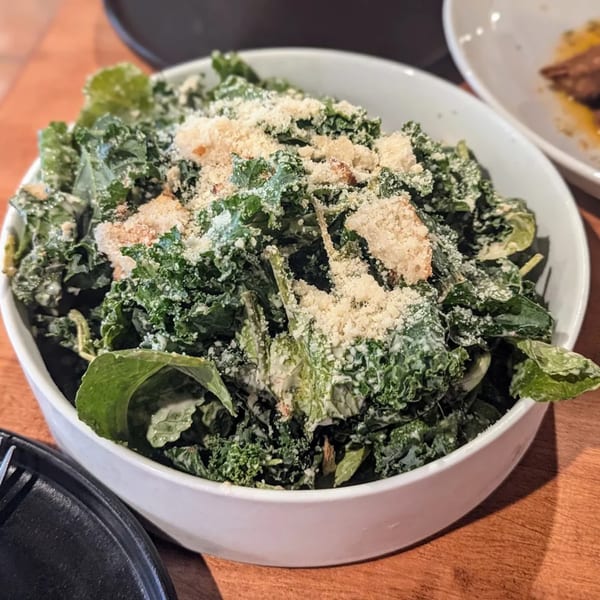 A white bowl of kale Caesar salad with croutons and parmesan cheese on a wooden table.