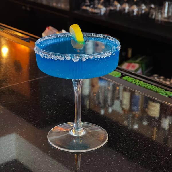 A vibrant blue cocktail with a salted rim and lemon garnish sits on a dark bar.