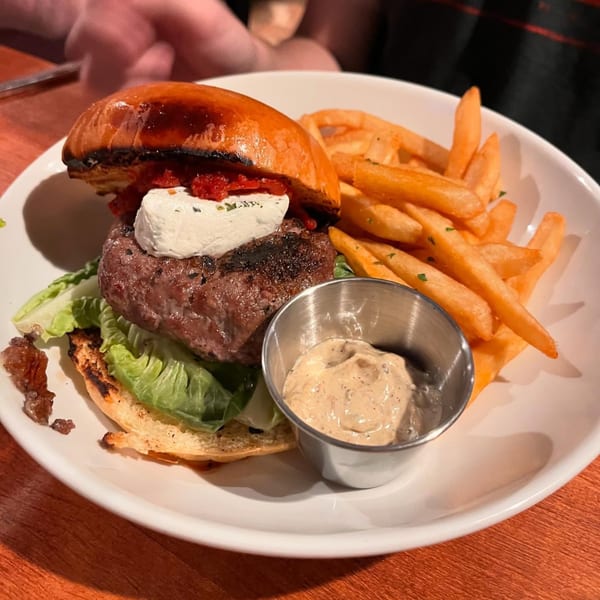A gourmet burger with goat cheese, roasted red peppers, and lettuce, served with fries and dipping sauce.