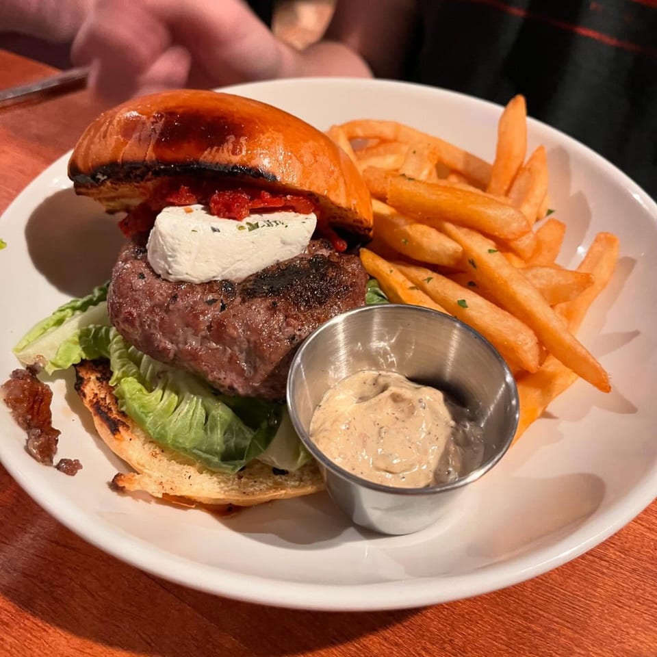A gourmet burger with goat cheese, roasted red peppers, and lettuce, served with fries and dipping sauce.