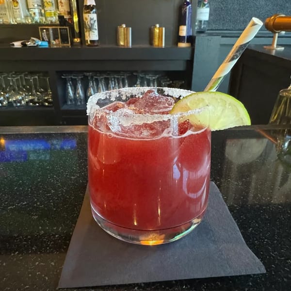A vibrant red cocktail with a salted rim, lime wedge, and striped straw on a dark bar.